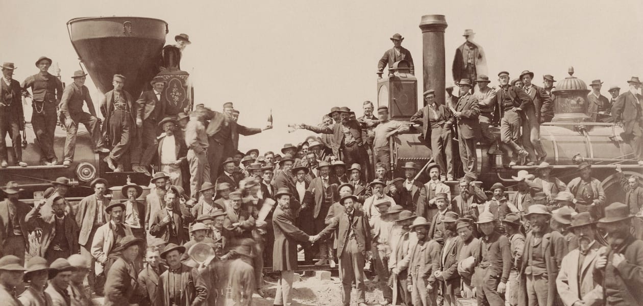 Railroad crews crowd onto two steam locomotives meeting nose to nose at Promontory Summit in 1869, as two men shake hands where the last rail was laid